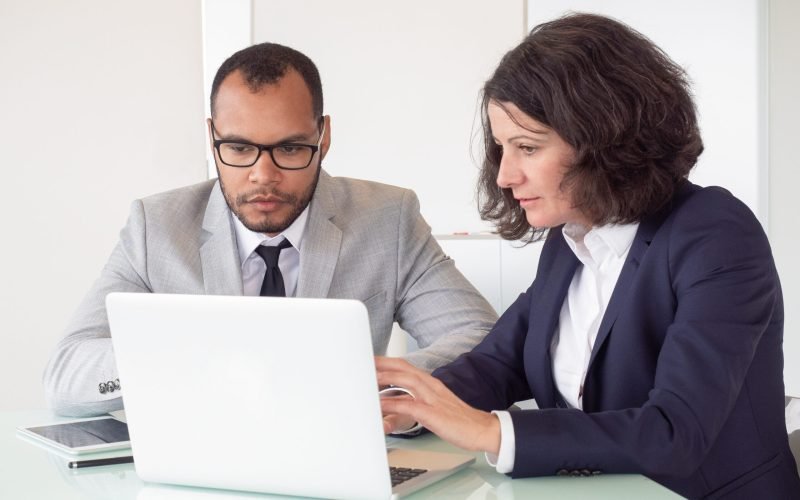 Serious coworkers using laptop. Professional multiethnic businessman and businesswoman sitting at table and working with laptop computer in office. Business and technology concept